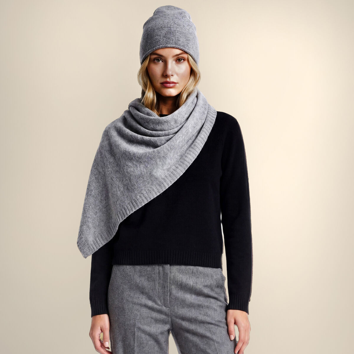 Eine Frau trägt die Kaschmirstola "Donna" Graumelange locker um die Schultern, dazu eine passende graue Mütze, schwarzen Pullover und graue, weite Stoffhose. Die Szenerie wirkt elegant und stilvoll, der Hintergrund ist neutral beige und lenkt nicht ab.