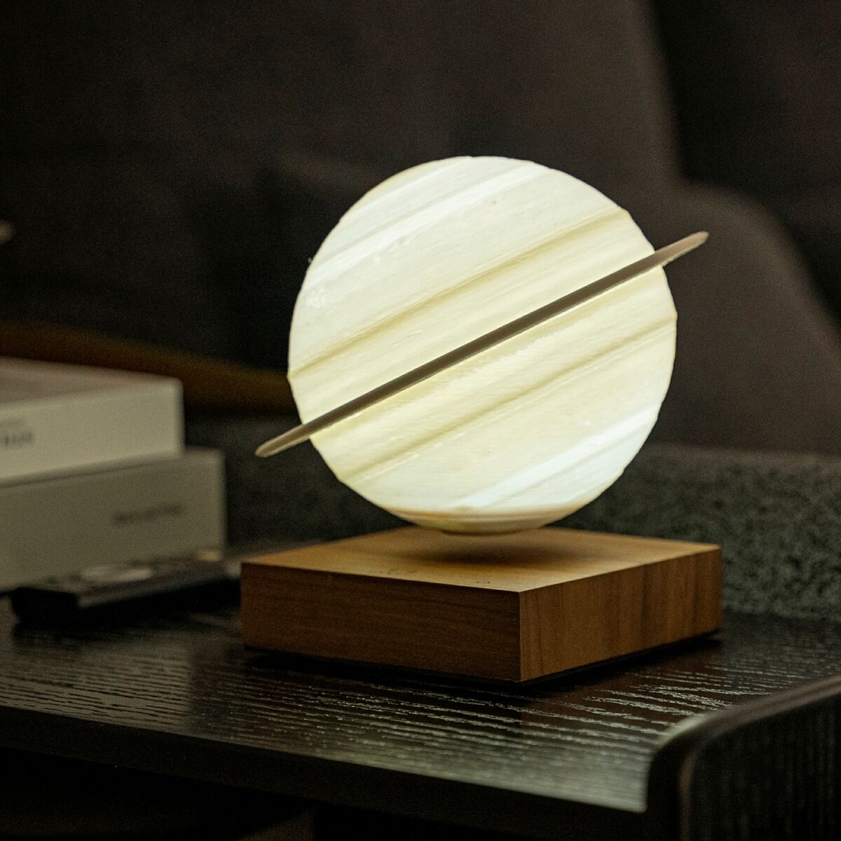 Schwebende Lampe in Form von Saturn, mit Holzsockel und LED-Beleuchtung auf einem Tisch, umgeben von Büchern, im Wohnzimmer.