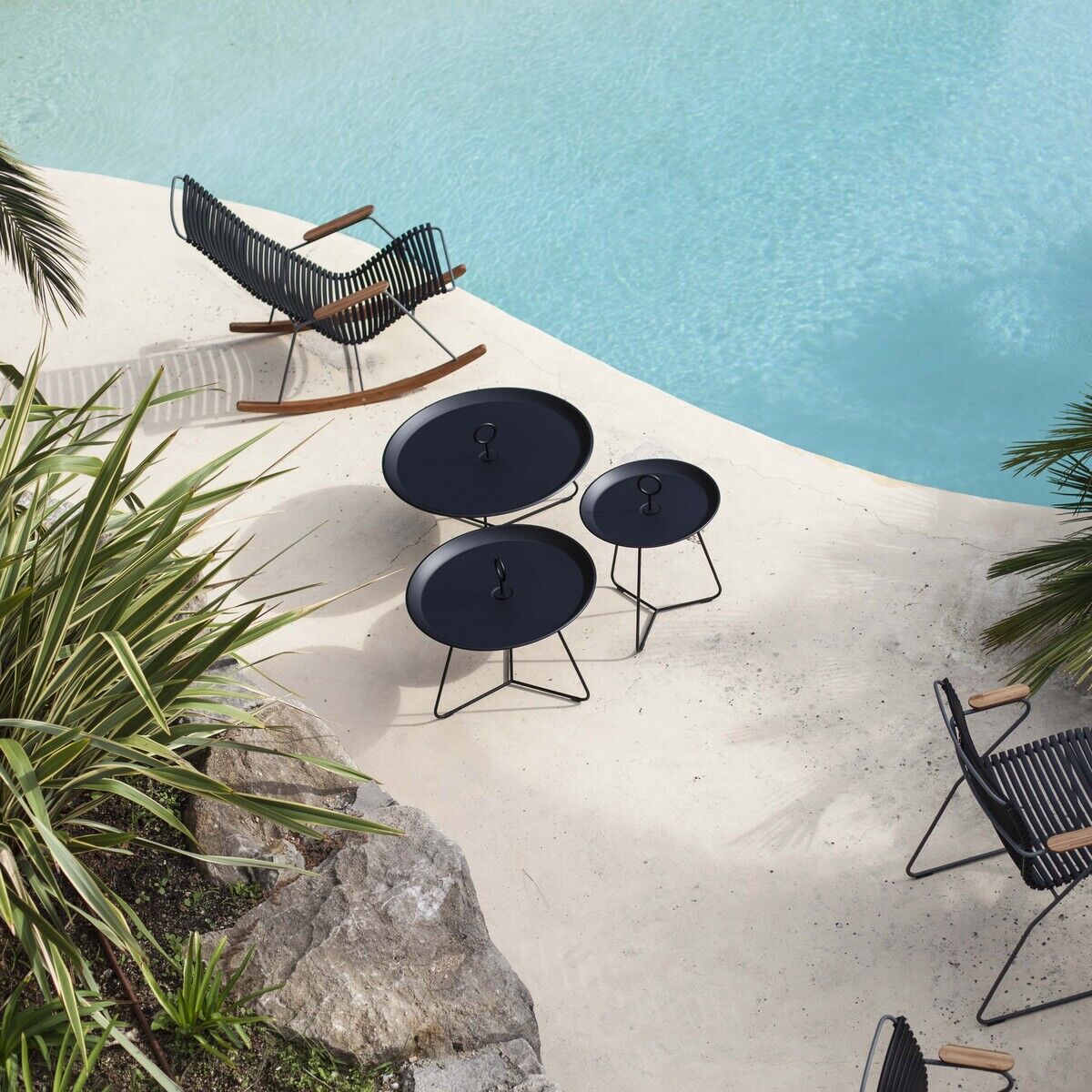 Ein Bild zeigt mehrere Beistelltische der Serie »EYELET« auf einer stilvollen Terrasse am Pool. Die Tische sind schwarz und rund mit einem minimalistisch modernen Design, umgeben von verschiedenen Pflanzen und Gartenmöbeln.