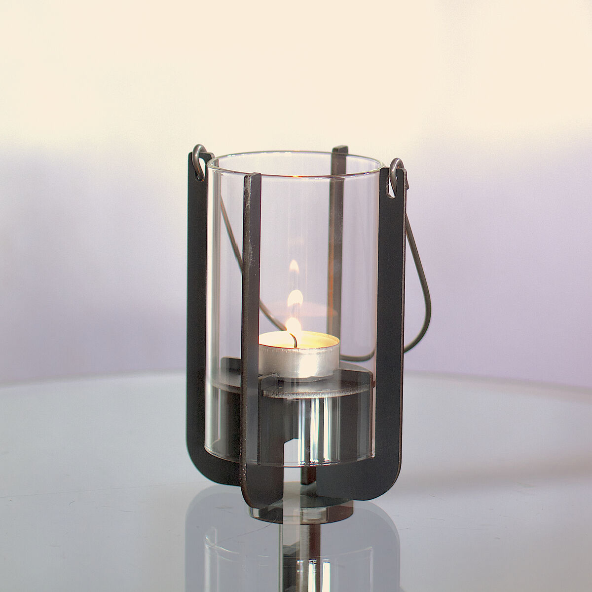 Eine moderne Gartenlaterne namens 'Lumi' steht auf einem Glas-Beistelltisch. Die Laterne ist aus schwarzem Metall und Glas gefertigt und enthält eine brennende Kerze. Im Hintergrund steht ein rostroter Sessel vor einer weißen Wand.