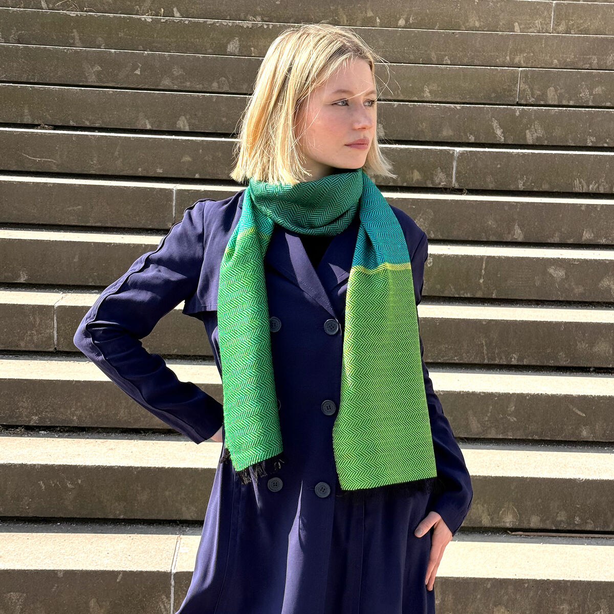Person trägt einen eleganten Seidenschal im Fischgrätmuster in den Farben Grün und Blau, der um den Hals gewickelt ist. Im Hintergrund sind Stufen zu erkennen.