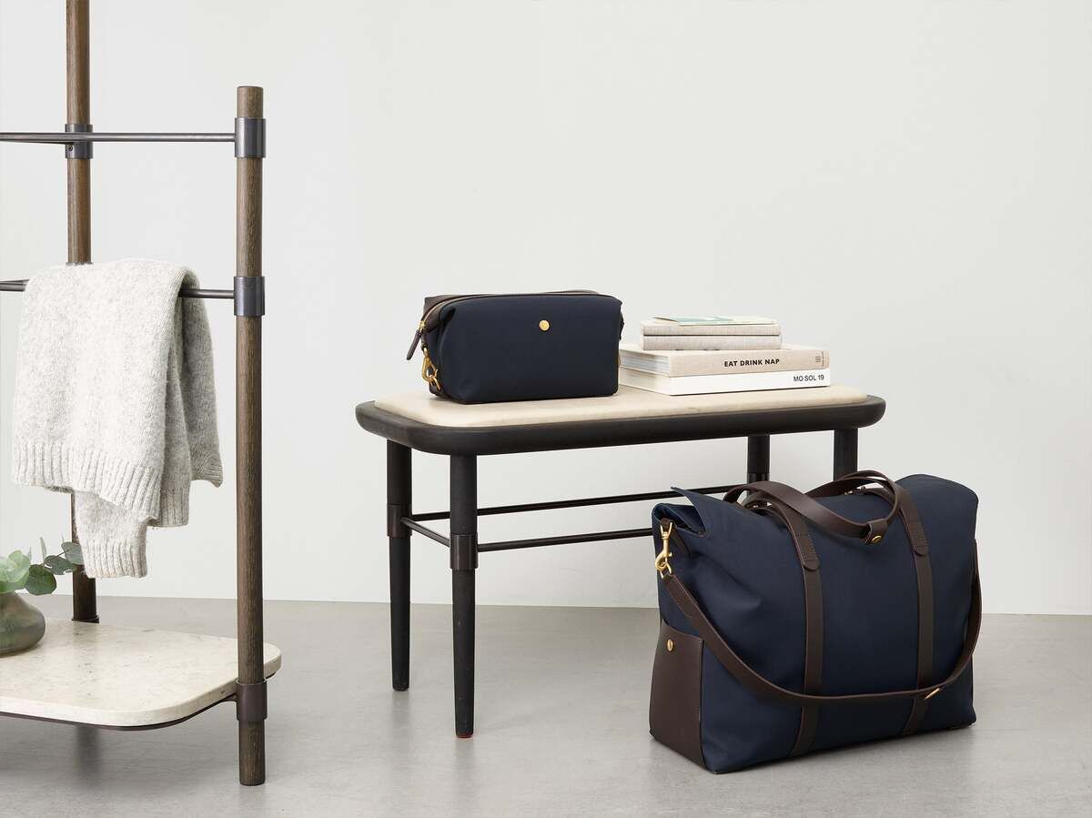 Eine elegante dunkelblaue Kulturtasche von Mismo zusammen mit einer passenden Reisetasche auf einem Tisch. Daneben Bücher und ein Handtuch auf einem Handtuchständer.