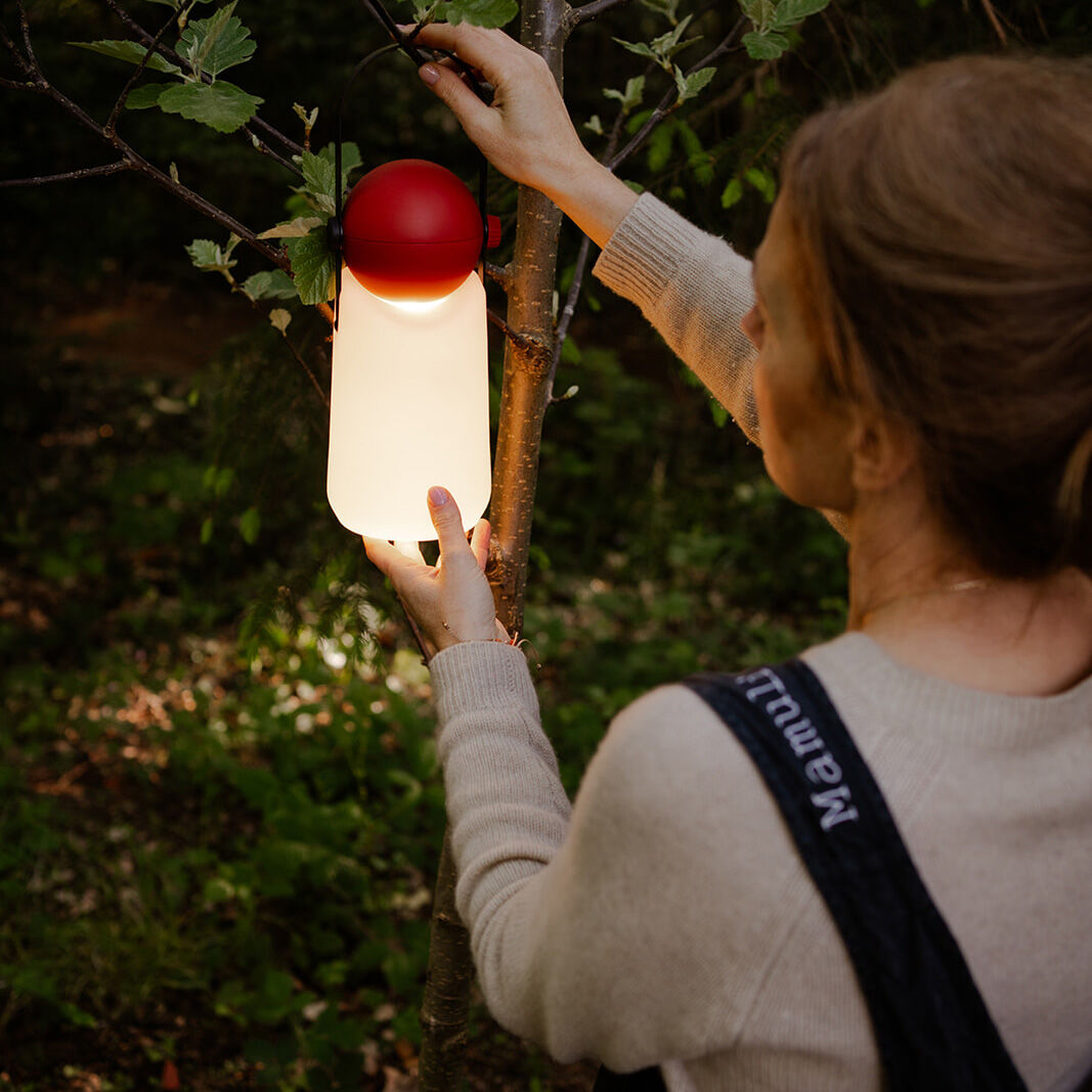 Eine Frau hängt die tragbare Leuchte "Guidelight" der Marke Weltevree mit rotem Kopf und weißem Leuchtkörper an einen Ast. Sie steht im Garten, umgeben von grünem Laub und Baumstämmen. Die Lampe leuchtet warm in der Dämmerung und sorgt für gemütliche Atmosphäre.