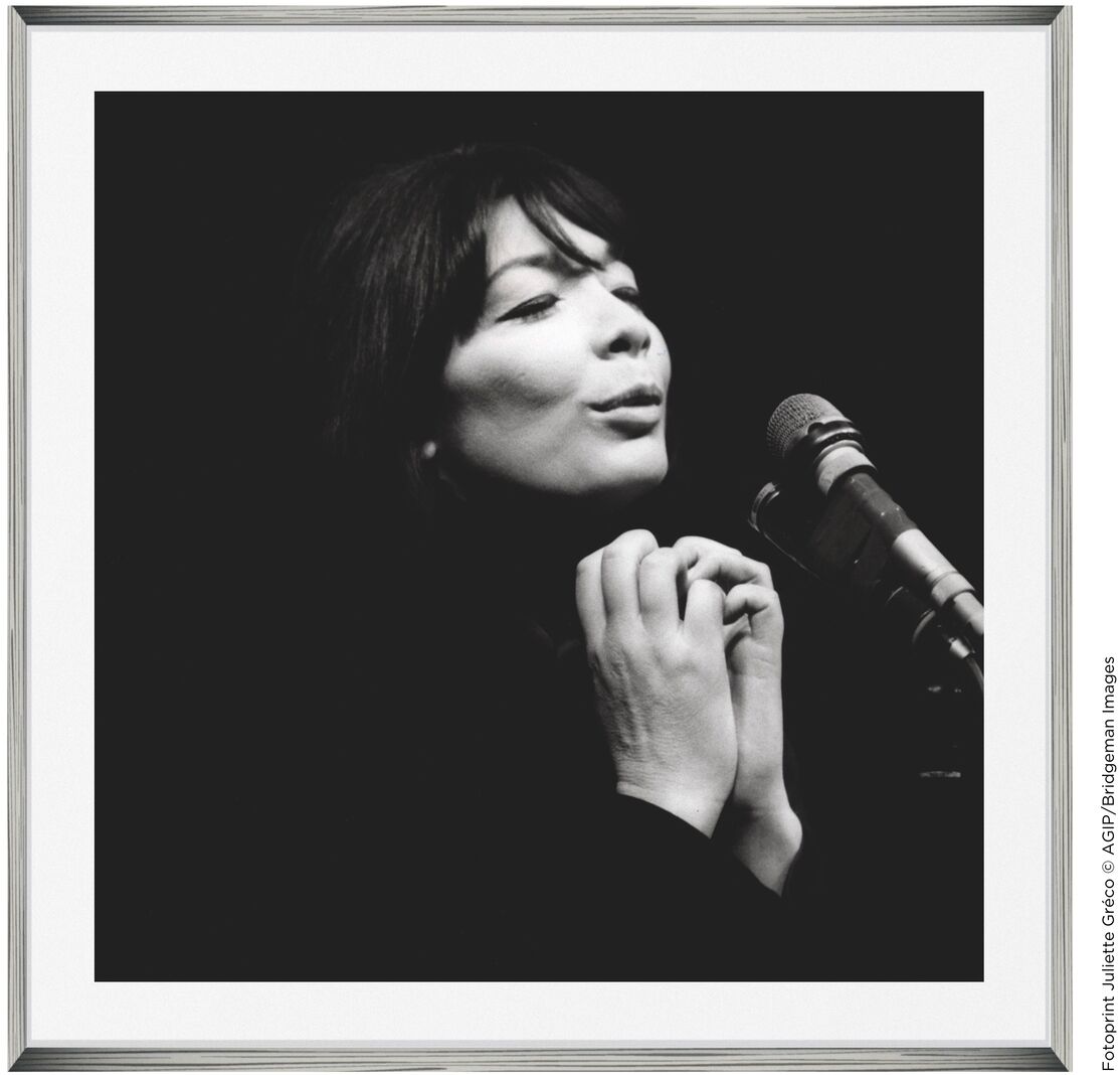 Schwarz-weiß Fotografie einer Sängerin, die mit geschlossenen Augen in ein Mikrofon singt. Die Hände sind nahe am Gesicht gefaltet. Der Rahmen ist schlicht und rechteckig.