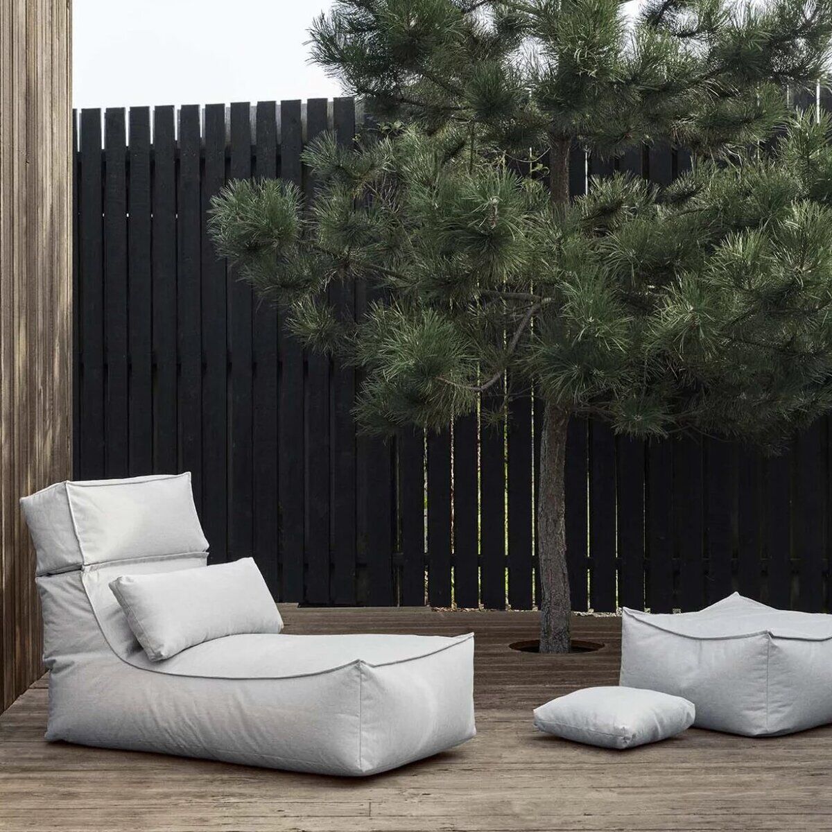 Ein Outdoor-Lounger-Set »STAY«, bestehend aus grauen Sitzgelegenheiten auf einem hölzernen Terrassenboden, mit einem schwarzen Holzzaun und einem grünen Baum im Hintergrund.