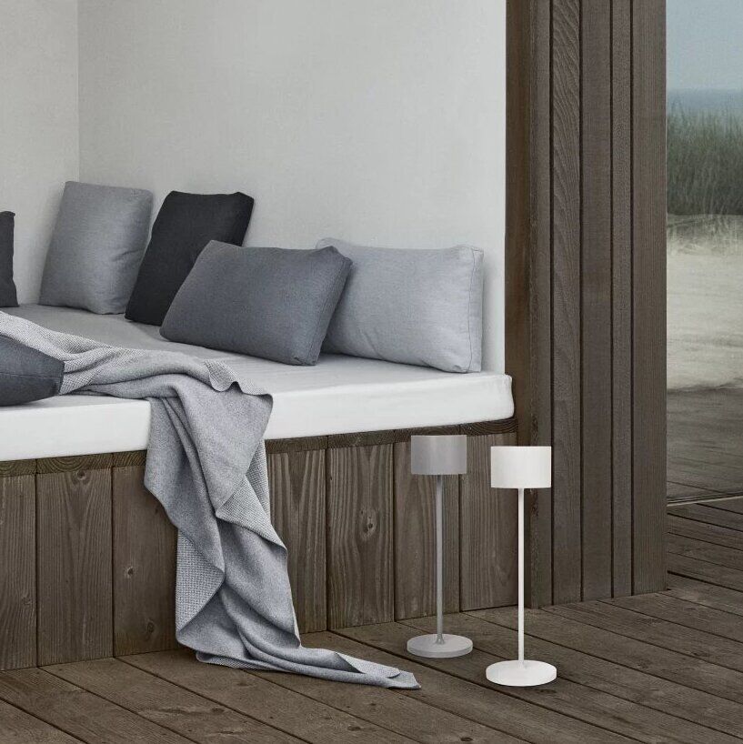Eine moderne, kabellose LED-Tischleuchte »FAROL« steht auf einem Holztisch neben einem gemütlichen Sitznischenbereich mit Kissen und Decken in einem minimalistischen Außenbereich eines Holzhauses.