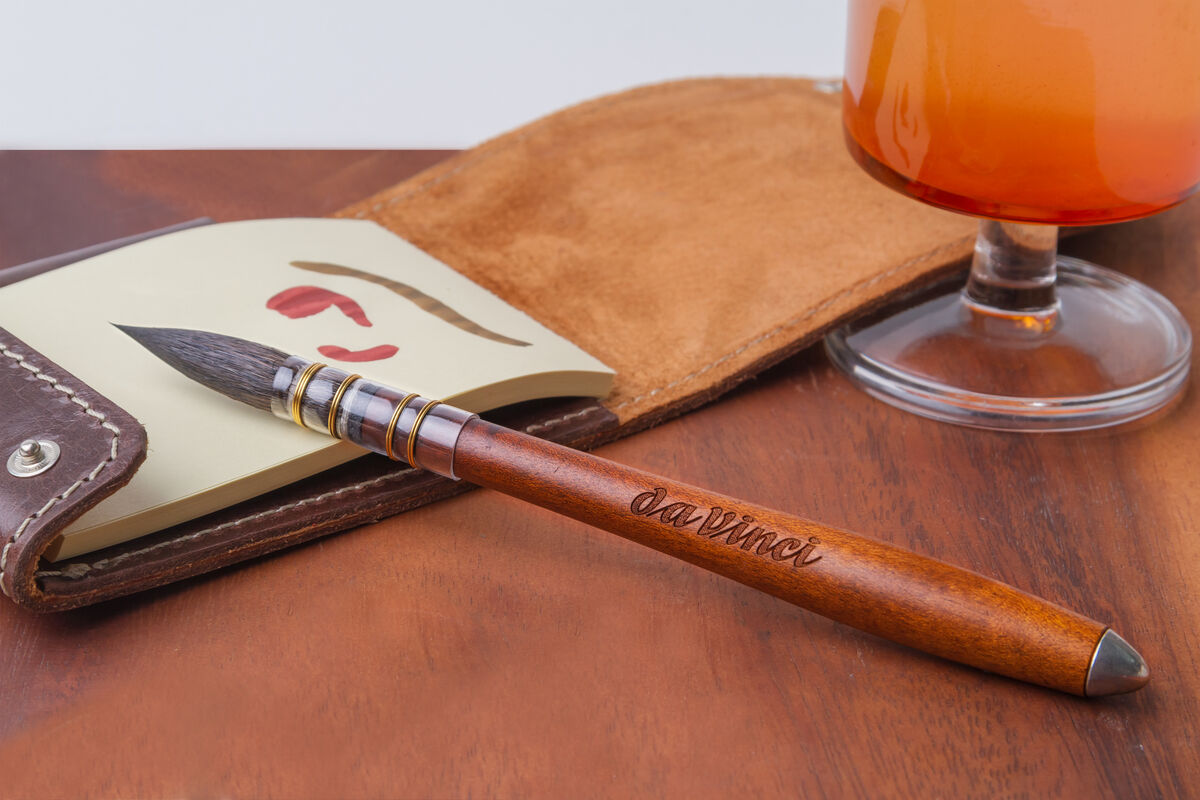 Traditioneller Aquarellpinsel mit Ethergraf®-Metallspitze, liegend auf einem Holztisch neben einer geöffneten ledernen Pinseltasche und einem Glas mit einem roten Getränk.