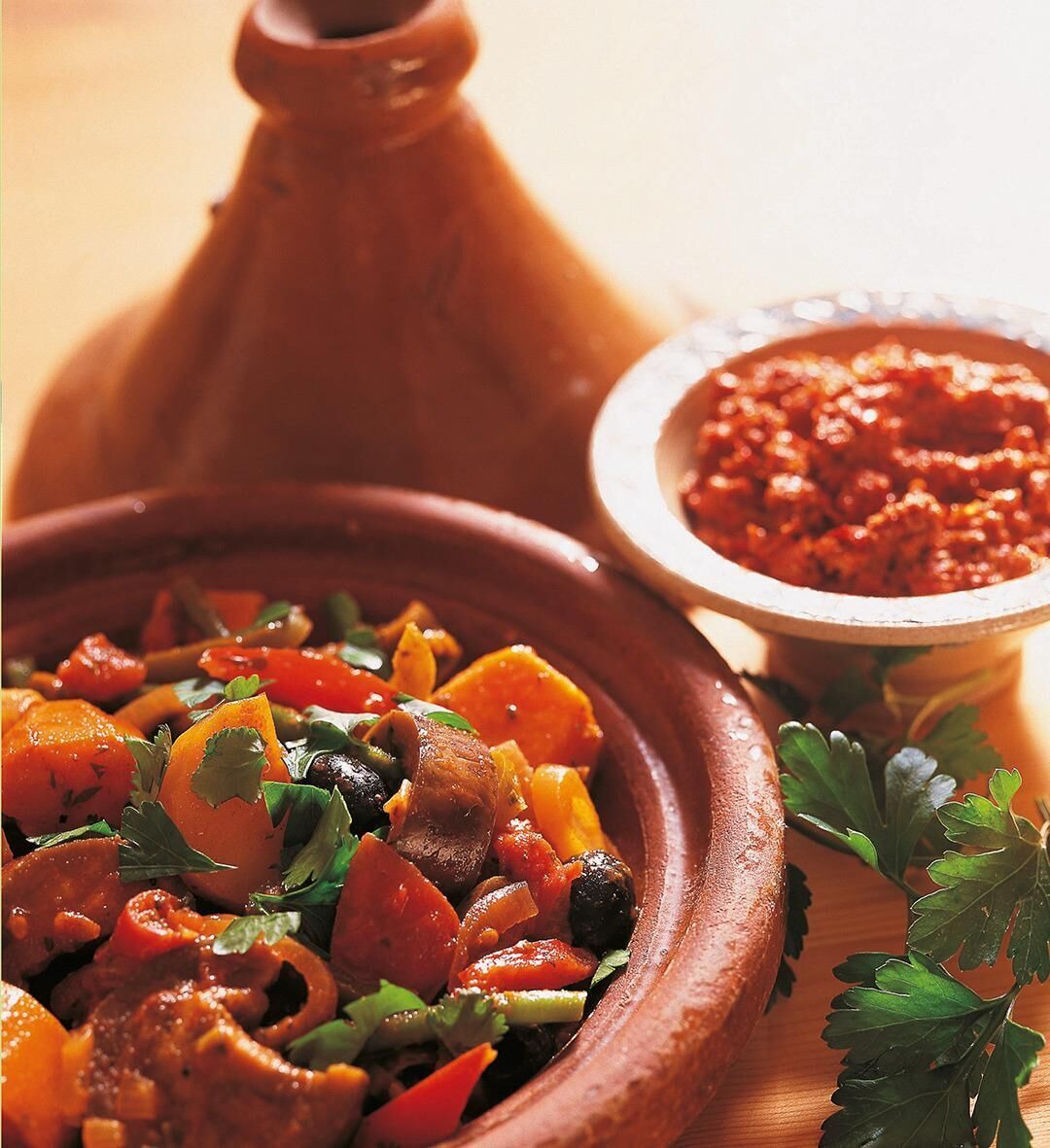 Ein Arrangement aus leckerem vegetarischem Essen in einer Terrakotta-Schüssel, garniert mit frischen Kräutern, daneben ein kleiner Topf mit einer roten Soße auf einem hölzernen Tisch. Im Hintergrund steht eine Tajine.