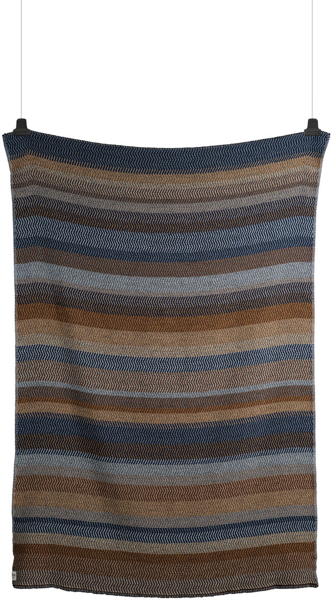 Lammwolldecke »Fri« mit Streifenmuster in verschiedenen Blau-, Braun-, Beige- und Grautönen, aufgehängt vertikal an zwei schwarzen Haken.