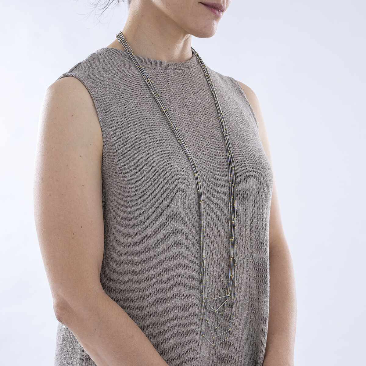Eine Frau trägt eine lange, graue und silberne Halskette über einem ärmellosen, grauen Kleid. Die Kette ist mehrlagig und besitzt einen minimalistischen Stil.