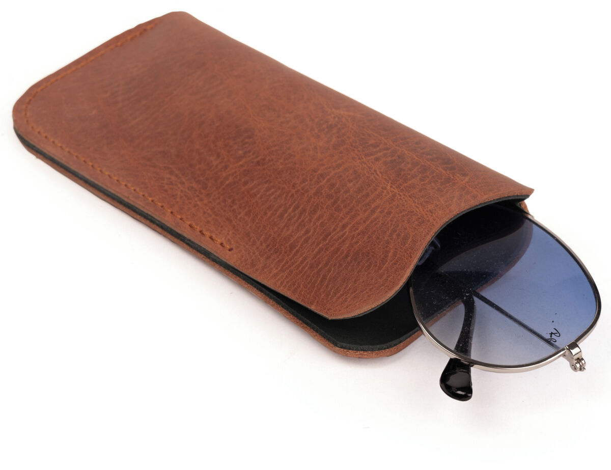 Auf weißem Hintergrund liegt ein hochwertiges, rechteckiges Brillenetui aus cognacfarbenem Kernleder. Aus dem geöffneten Etui ragt eine Sonnenbrille mit dunkelblauen Gläsern und schwarzem Gestell. Das Leder zeigt eine feine Maserung.