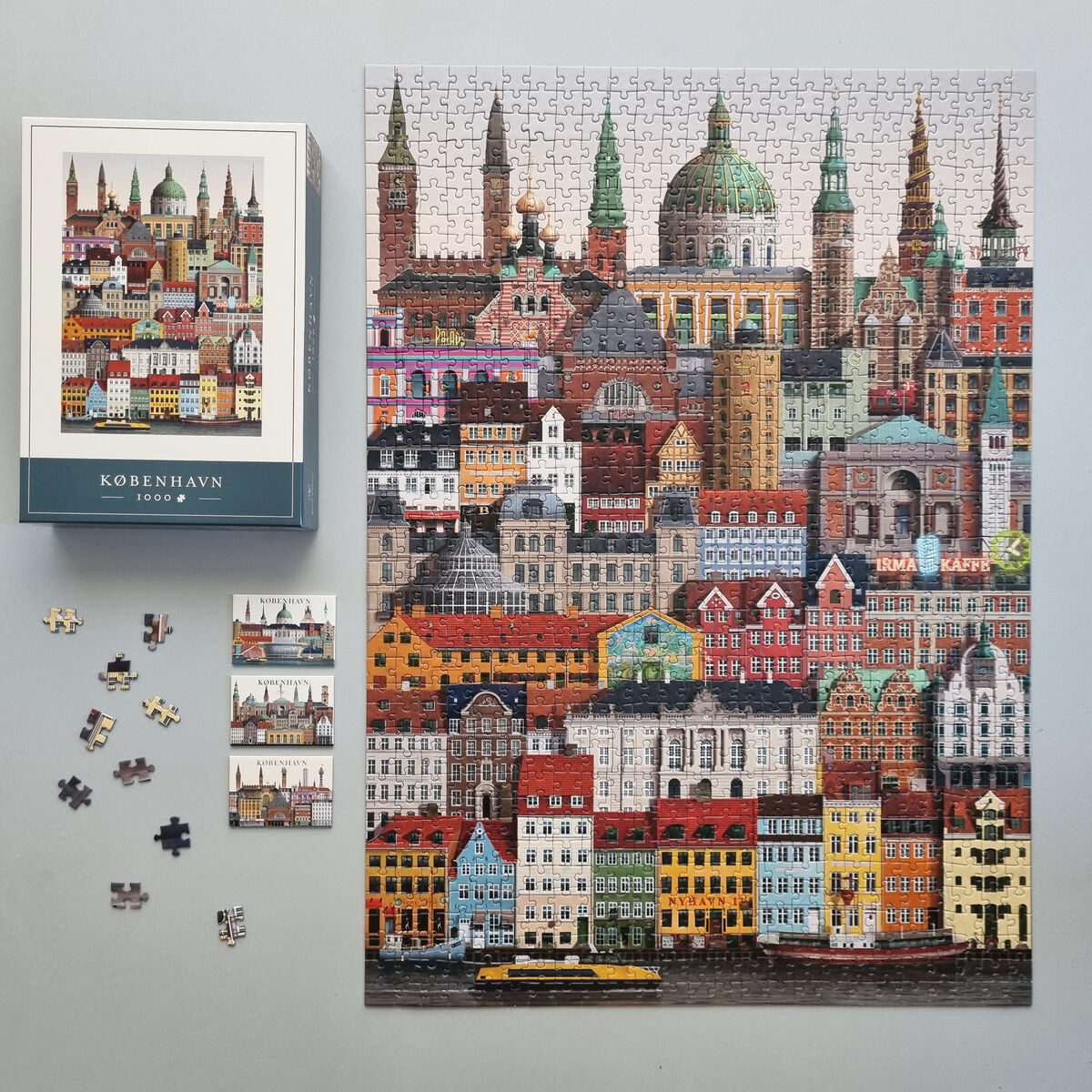 Das Bild zeigt ein fertiges 1000 Teile Puzzle mit einer detaillierten Stadtansicht von Kopenhagen, das viele bunte Gebäude und markante Architektur zeigt. Neben dem Puzzle liegt die Originalverpackung und einige noch nicht gelegte Puzzleteile. Der Hintergrund ist neutral grau gehalten, wodurch die leuchtenden Farben der Gebäude hervorgehoben werden.