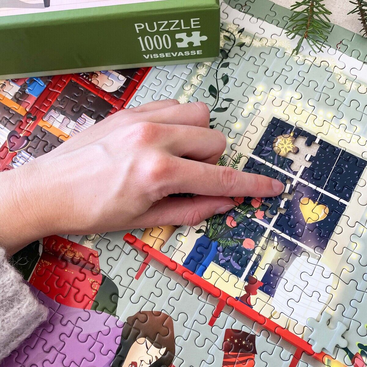 Das Bild zeigt ein fast fertiggestelltes 1000-Teile Puzzle von VISSEVASSE mit winterlich-weihnachtlichem Motiv. Eine Hand legt das letzte Teil in ein Fenster mit Schneeflocken. Im Vordergrund ist eine Verpackung mit der Aufschrift „Puzzle 1000 VISSEVASSE“ zu sehen.