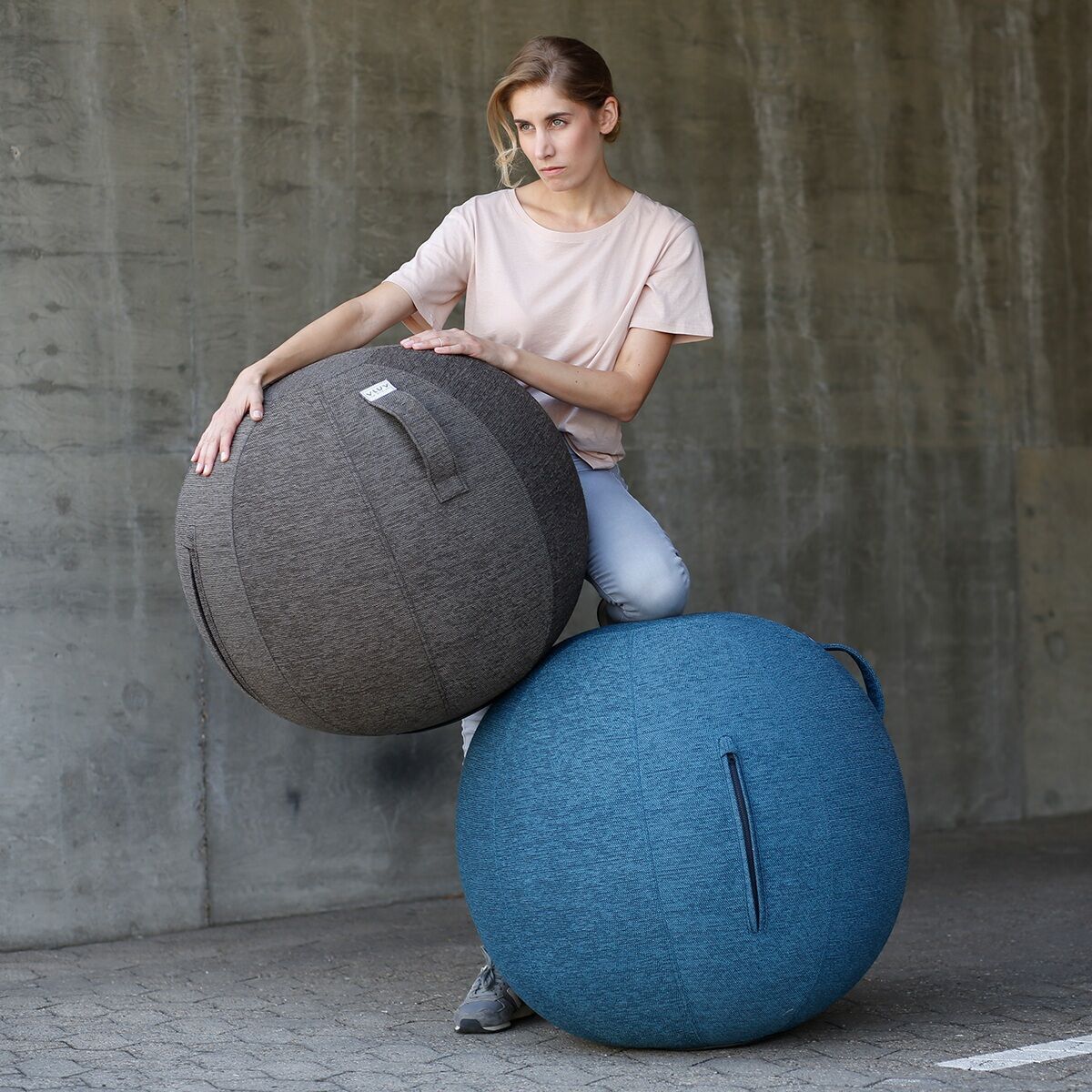 Eine Frau sitzt auf einem petrolfarbenen Sitzball »Vluv Stov« von HOCK Design und hält einen weiteren Sitzball in Anthrazit. Beide Bälle bestehen aus grobem Polyesterstoff und haben einen praktischen Tragegriff. Im Hintergrund befindet sich eine graue Betonwand.