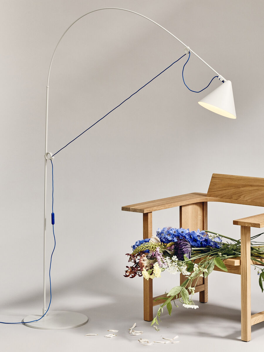 Eine modern gestaltete Stehlampe der ZEIT-Sonderedition AYNO in den Farben Silk und Ultramarin steht neben einem hellen Holzstuhl. Auf dem Stuhl liegt ein bunter Blumenstrauß. Die Szene wirkt minimalistisch und elegant, mit neutralem Hintergrund und zurückhaltender Farbgestaltung.