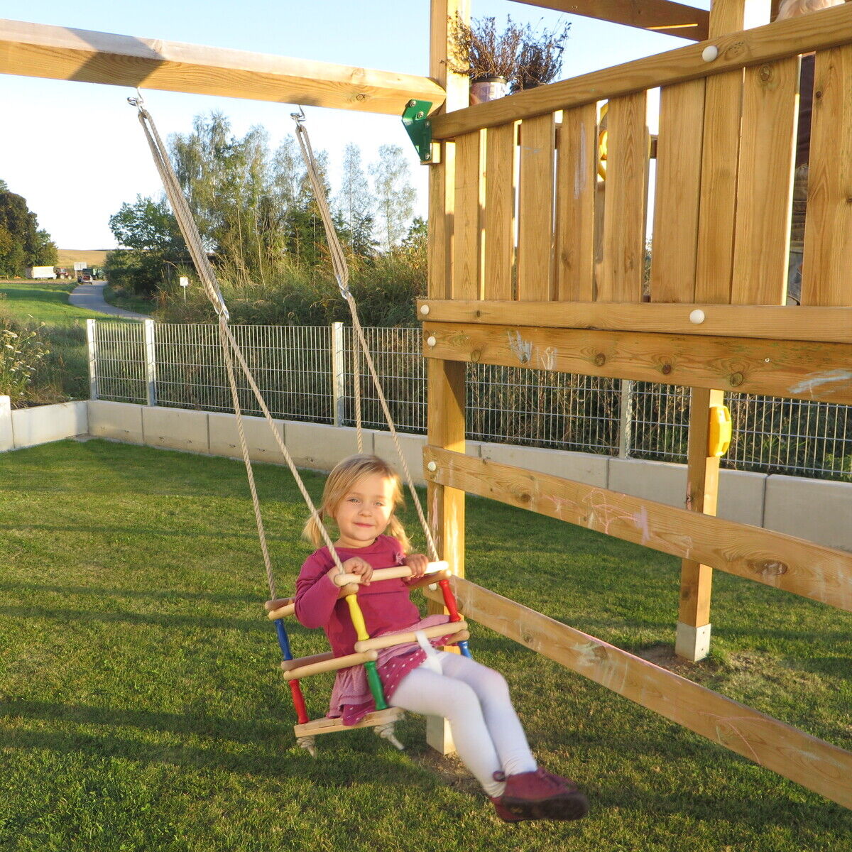 Ein kleines Mädchen sitzt auf einer Schaukel aus Buchenholz, die an einem hölzernen Spielgerüst im Garten befestigt ist. Im Hintergrund sind Gras, ein Zaun und Bäume zu sehen. Das Kind trägt ein rotes Oberteil und weiße Strumpfhosen.