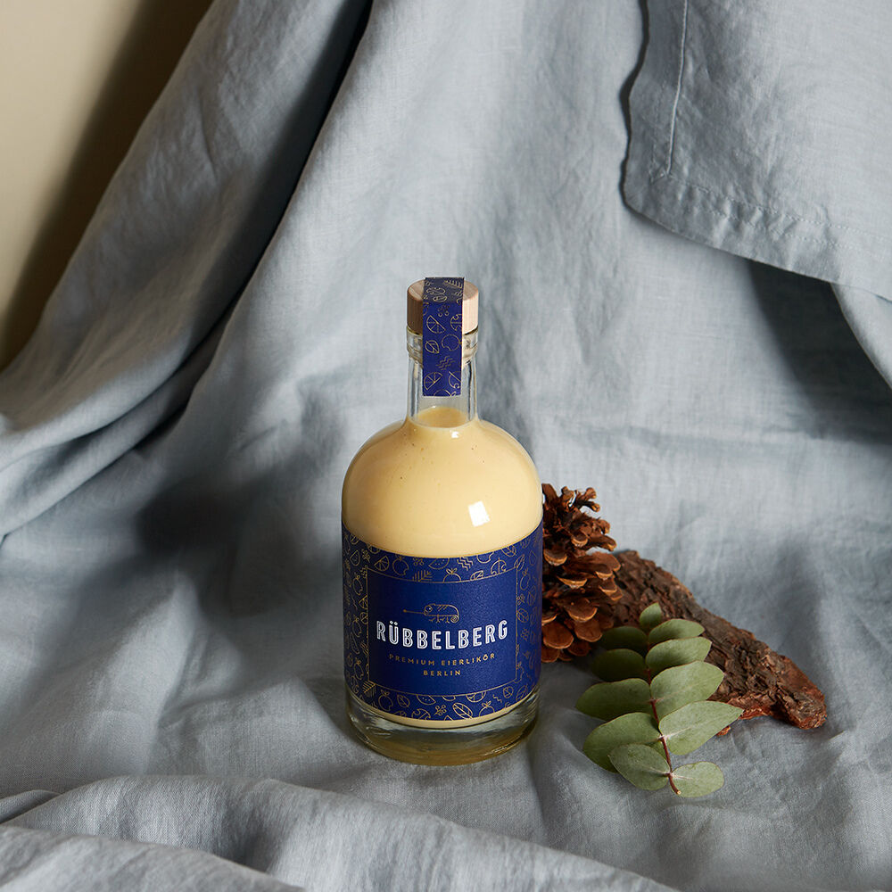 Eine Flasche »Premium Eierlikör« von Rübbelberg mit cremefarbener Likörfüllung und edlem, dunkelblauem Etikett steht auf hellblauem Stoff. Daneben liegen eine Kieferzapfen und grüne Blätter. Das Arrangement wirkt modern und hochwertig.