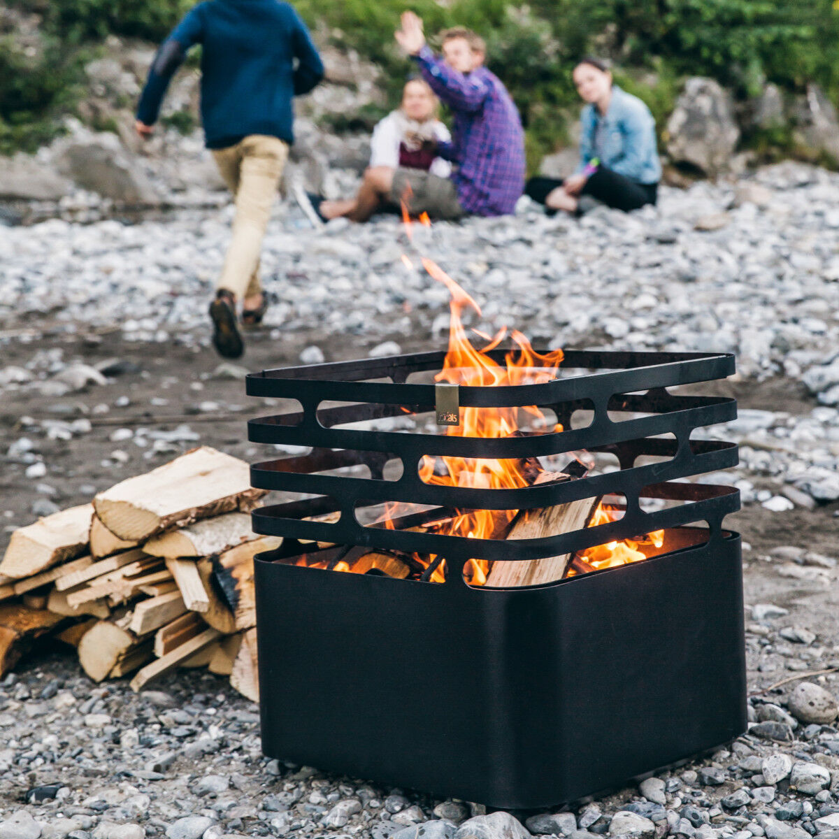 Eine moderne, quadratische Feuerstelle aus Metall mit brennendem Holz und Flammen darin, umgeben von Steinen, mit einem Stapel Holz daneben. Im Hintergrund sitzen vier Personen in Freizeitkleidung auf Steinen und unterhalten sich oder gehen.