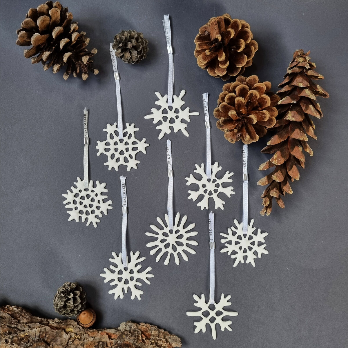Acht kleine Schneeflocken-Anhänger aus weißem Material mit Bändchen liegen auf dunkelgrauem Untergrund. Dazu sind vier große Tannenzapfen, zwei kleine Tannenzapfen, eine Rindenscheibe und eine Haselnuss dekorativ arrangiert. Die Schneeflocken stammen von Silke Decker.