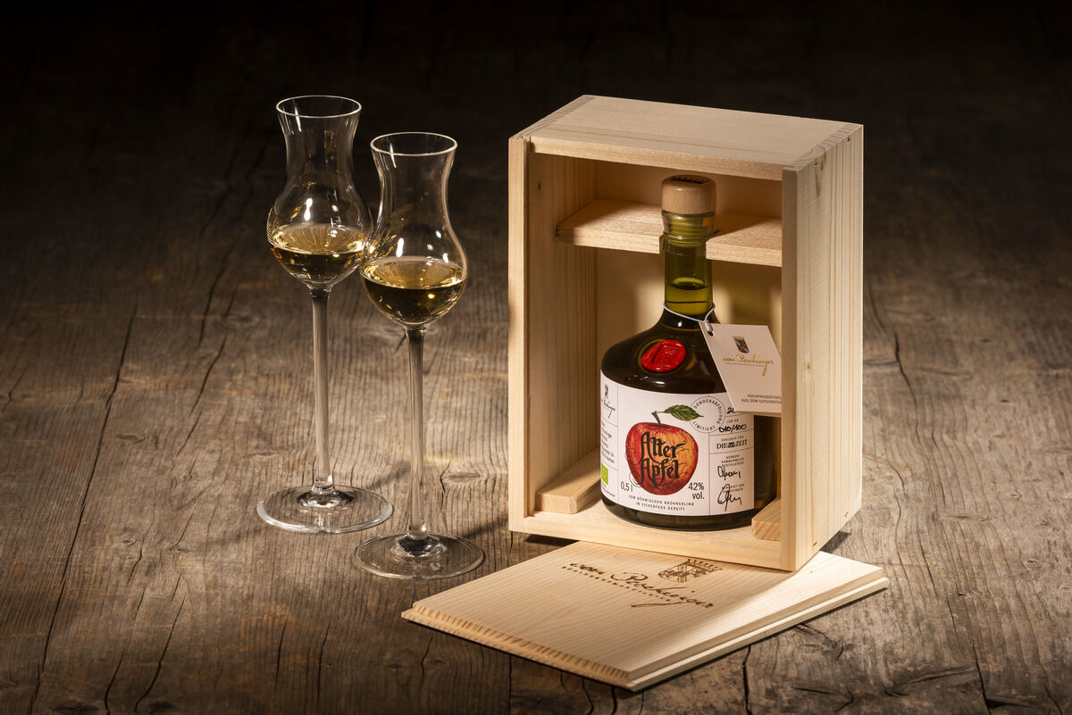 Eine Flasche ZEIT-Sonderedition Edelbrand »Alter Apfel« in einer hellen Holzkiste, begleitet von zwei halb gefüllten Schnapsgläsern und einem Notizbuch auf einem rustikalen Holztisch.