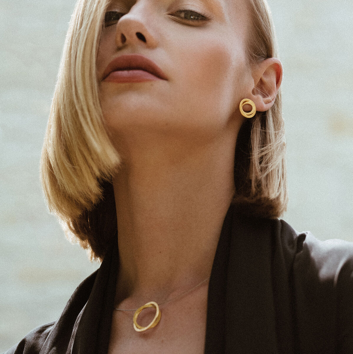 Eine Frau mit blonden Haaren trägt ein Collier mit einem großen, goldplattierten Silberanhänger. Sie trägt auch passende Ohrringe und einen schwarzen Blazer. Der Hintergrund ist verschwommen.