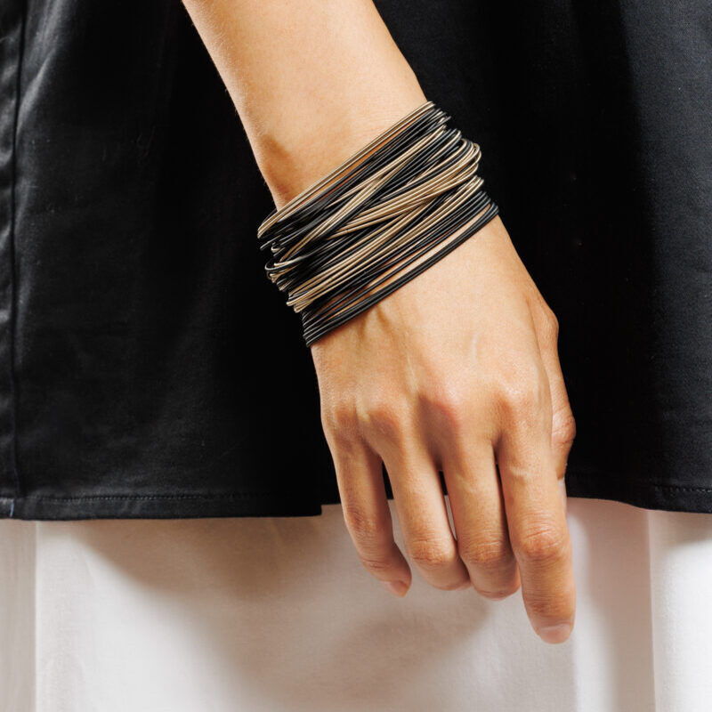 Detailreiches Bild eines Handgelenks mit einem modernen Armband-Set aus 60 elastischen Einzelbändern in Bronze und Schwarz von la molla. Das leichte Schmuckstück aus Chirurgenstahl liegt flexibel und anschmiegsam am Handgelenk auf. Der Untergrund ist schwarz-weiß gekleidet und hebt das Armband optisch hervor.