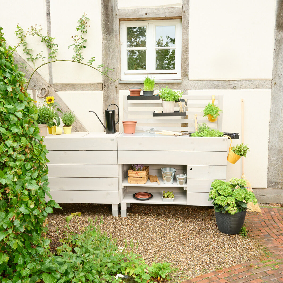 Eine Beetbar in einem Garten mit verschiedenen Pflanzen, Kräutern und Gartenwerkzeugen. Die Beetbar ist aus hellem Holz gefertigt und steht auf einem Kiesboden. Im Hintergrund ist ein weißes Haus mit Fachwerk zu sehen. Rechts daneben befindet sich weiteres Pflanzenzubehör in Töpfen.