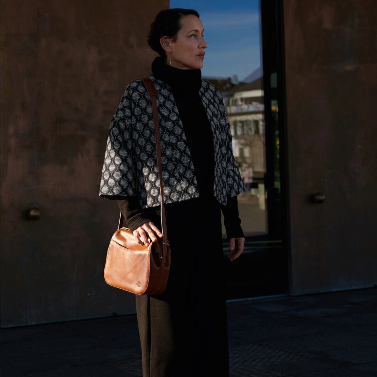 Frau trägt eine braune Ledertasche über der Schulter. Sie hat dunkle Kleidung an und steht vor einer urbanen Kulisse. Die Kombination aus der braunen Tasche und ihrer Kleidung strahlt Stil und Eleganz aus.