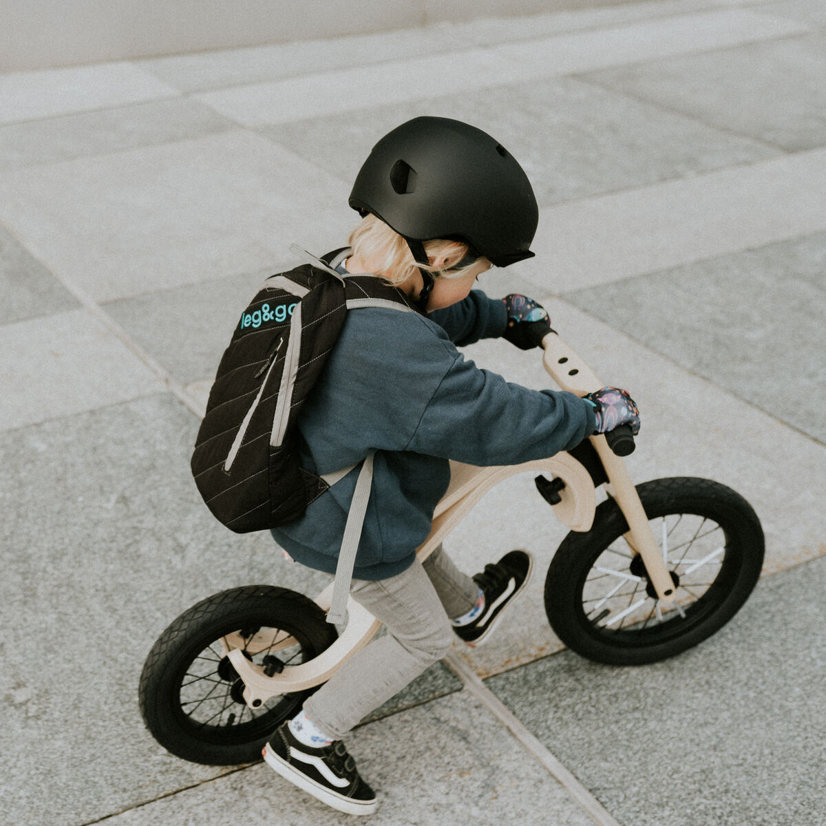 Ein kleines Kind mit Helm fährt auf einem hellen 3-in-1-Laufrad auf einem gepflasterten Platz. Das Kind trägt einen Rucksack und sportliche Kleidung. Die Szene vermittelt Bewegung und Freude in einem städtischen Außenbereich.