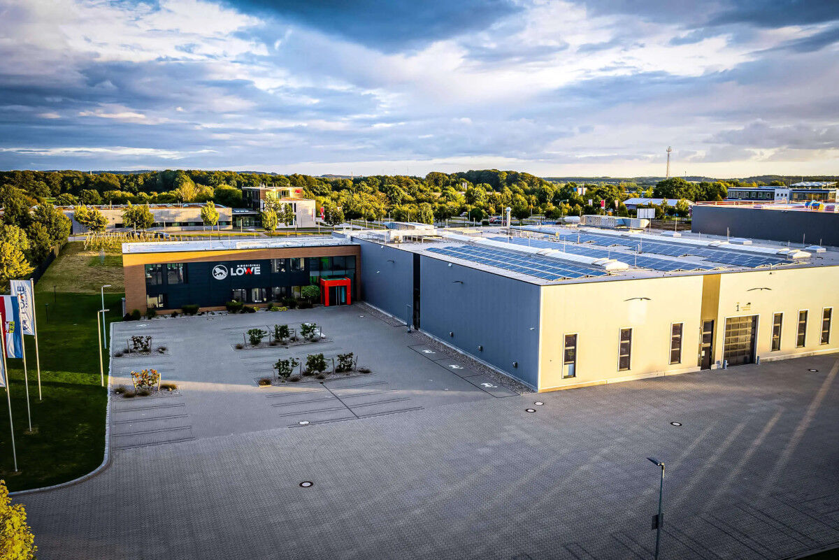 Luftaufnahme eines modernen Firmengebäudes mit Solaranlagen auf dem Dach und gepflastertem Vorplatz, umgeben von Bäumen.