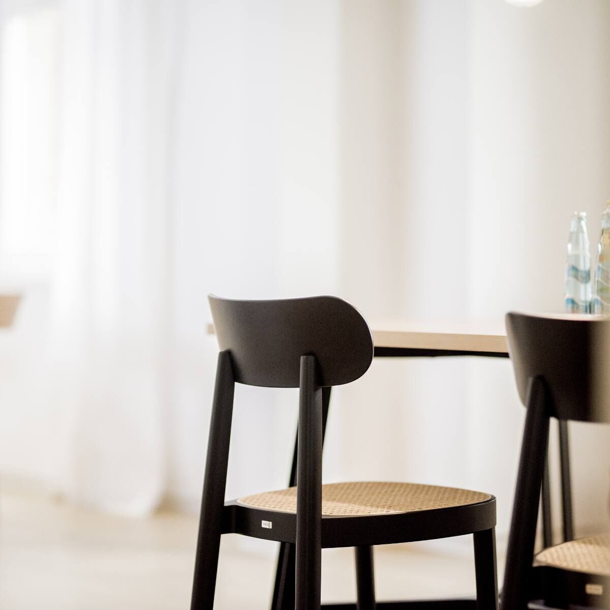 Ein minimalistischer Holzstuhl des Modells '118' von Thonet. Der Stuhl hat ein schlichtes Design und besteht aus schwarzem Holz mit einer beigen Sitzfläche. Er steht in einem hellen Raum mit weißen Vorhängen und einem Tisch im Hintergrund.