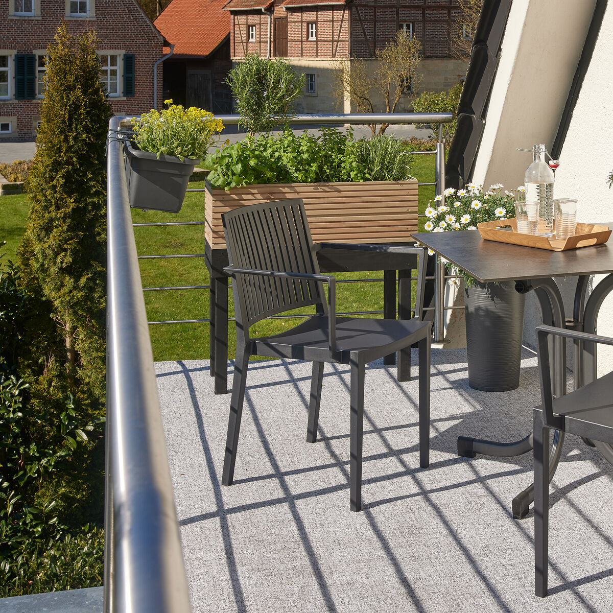 Ein Balkon mit einem Hochbeet »Nola« und zwei grauen Stühlen neben einem Tisch. Das Hochbeet ist mit grünen Pflanzen gefüllt. Im Hintergrund ist eine grüne Rasenfläche und das Gebäude zu sehen.