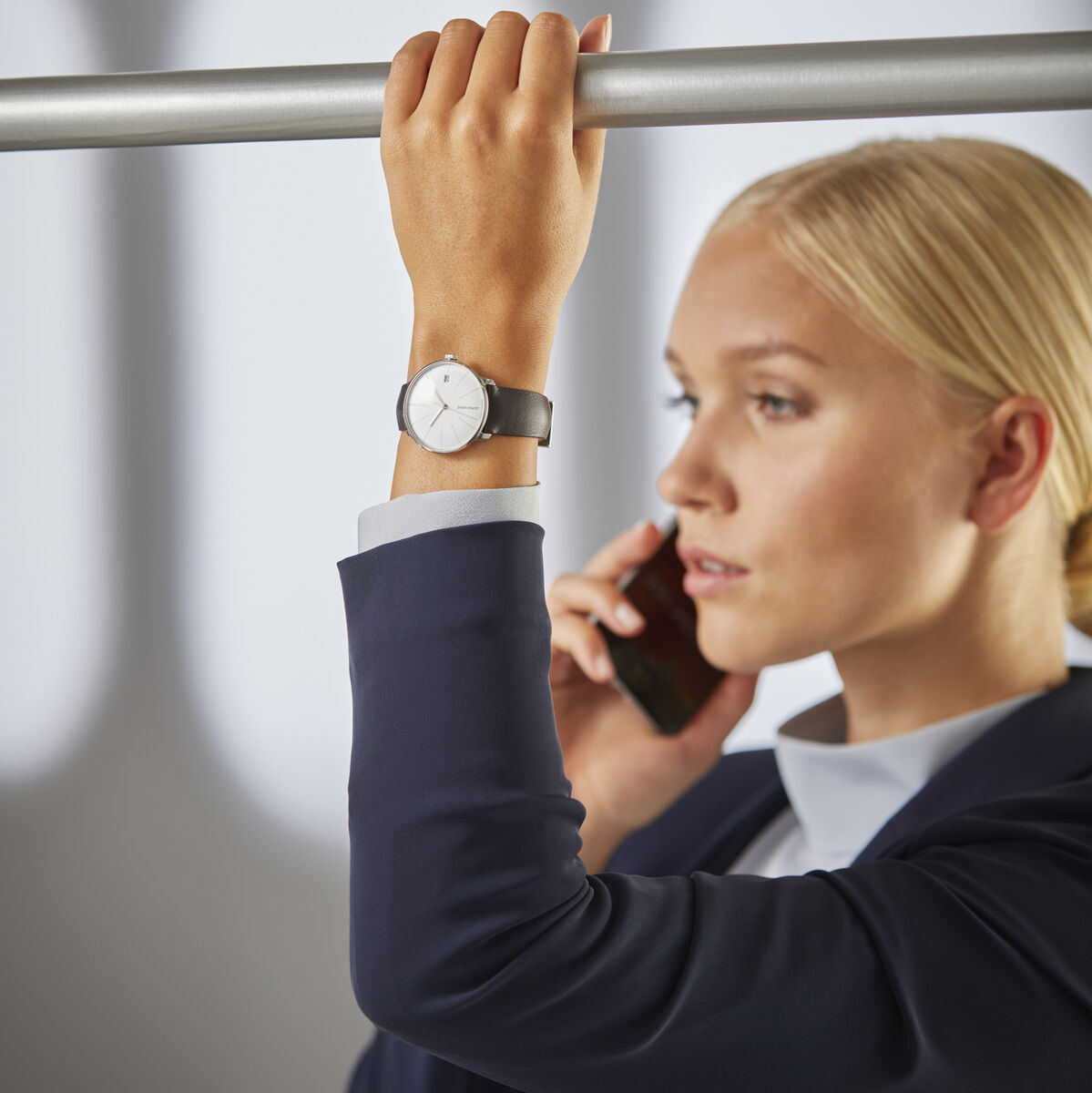 Eine Frau in Business-Kleidung hält sich in einem Transportmittel fest und telefoniert. Am Handgelenk trägt sie die ZEIT-Sonderedition 'Meister fein Kleine Automatic' von Junghans mit einem schwarzem Lederarmband und einem minimalistischen weißen Ziffernblatt.