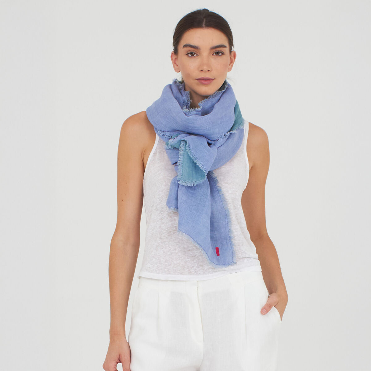 Eine Frau trägt den Leinenschal »Alsi« von Shalls Barcelona in Ocean. Der Schal aus 100% Leinen wirkt sehr leicht und weich, ist zweifarbig gefärbt und hat feine Fransen. Sie trägt ihn locker umgelegt zu einem weißen ärmellosen Top und weißen Hosen vor neutralem Hintergrund.