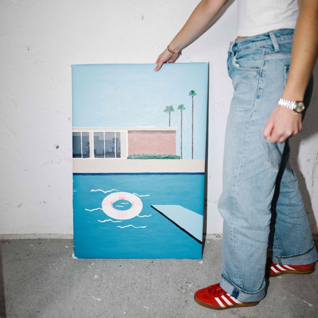 Eine Person in blauer Jeans und weißen T-Shirt hält ein Malen nach Zahlen Bild mit dem Motiv eines Pools und einer Gebäude im Hintergrund. Das Bild ist farbenfroh mit blauen und weißen Tönen.