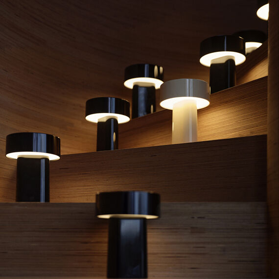 Mehrere tragbare Lampen vom Modell 'Oblong' in Schwarz und Weiß stehen beleuchtet auf einer Holztreppe in einer modernen, stilvoll eingerichteten Innenumgebung. Die Lampen sorgen für eine warme und gemütliche Lichtstimmung.