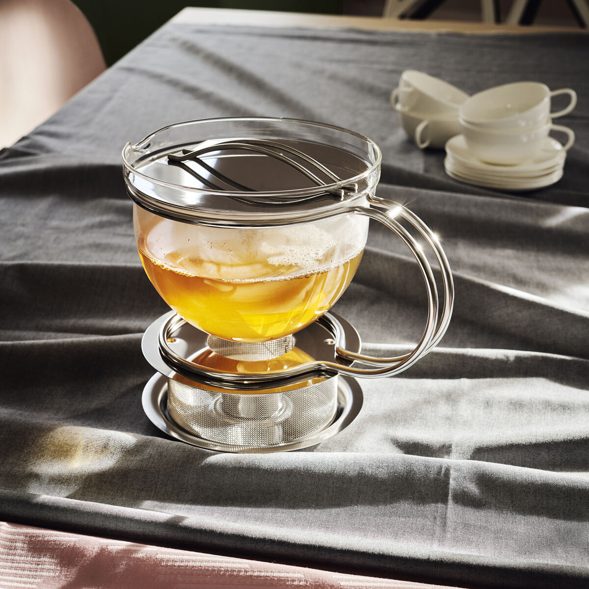 Eine moderne Teekanne »Filio« aus Glas und Metall steht auf einem gedeckten Tisch. Die Kanne ist mit goldgelbem Tee gefüllt und steht in einem silbernen Halter. Im Hintergrund sind gestapelte weiße Tassen zu sehen.