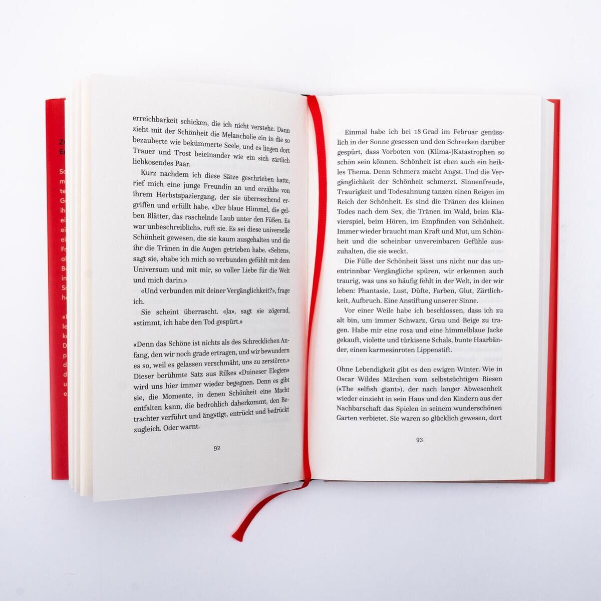 Ein geöffnetes Buch mit dem Titel 'Trost der Schönheit' von Arnim, G., das auf einer weißen Oberfläche liegt. Das Hardcover-Buch hat ein rotes Cover und ein rotes Leseband.