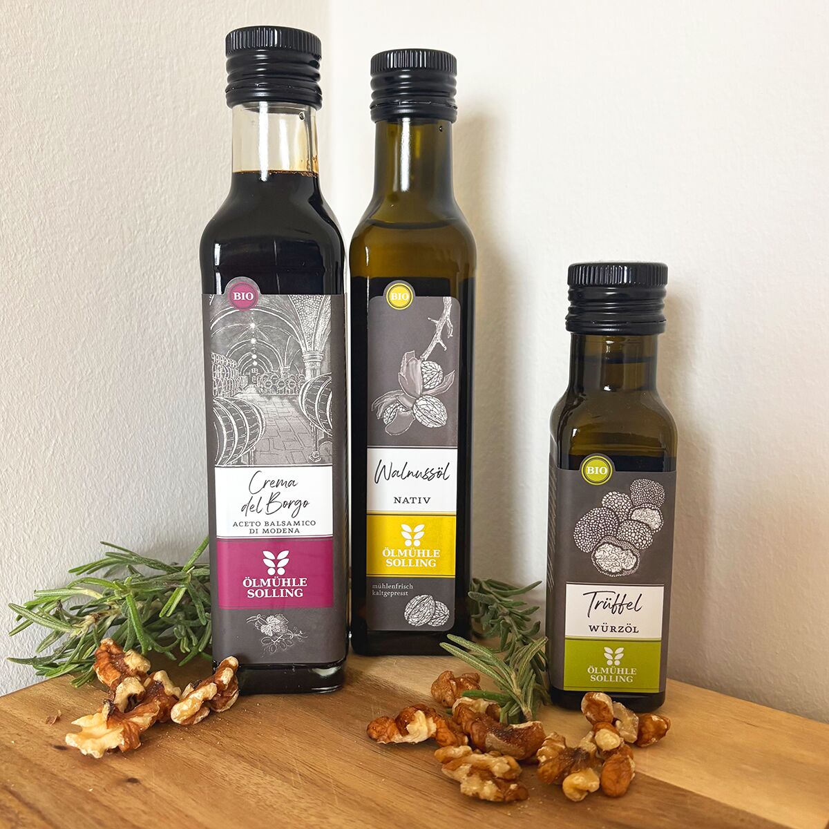 Drei Flaschen Bio-Öl (Crema di Balsamico, Walnussöl und Trüffelöl) der Marke Ölmühle Solling stehen nebeneinander auf einer Holzoberfläche. Dekoriert mit Rosmarinzweigen und Walnusshälften vor einer weißen Wand.