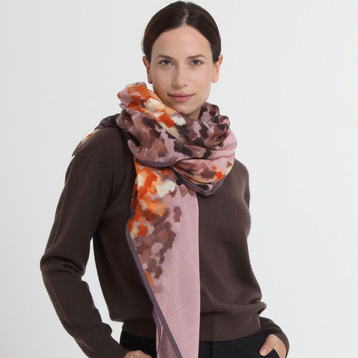 Frau mit elegantem Wollschal »Hazel« Mauve von Shalls Barcelona aus 100% Schurwolle. Der Schal ist mauvefarben mit abstraktem, buntem Muster in Rosa, Orange, Braun und Beige und großzügig um Hals und Schultern drapiert. Die Frau trägt einen braunen Pullover und schwarze Hose.