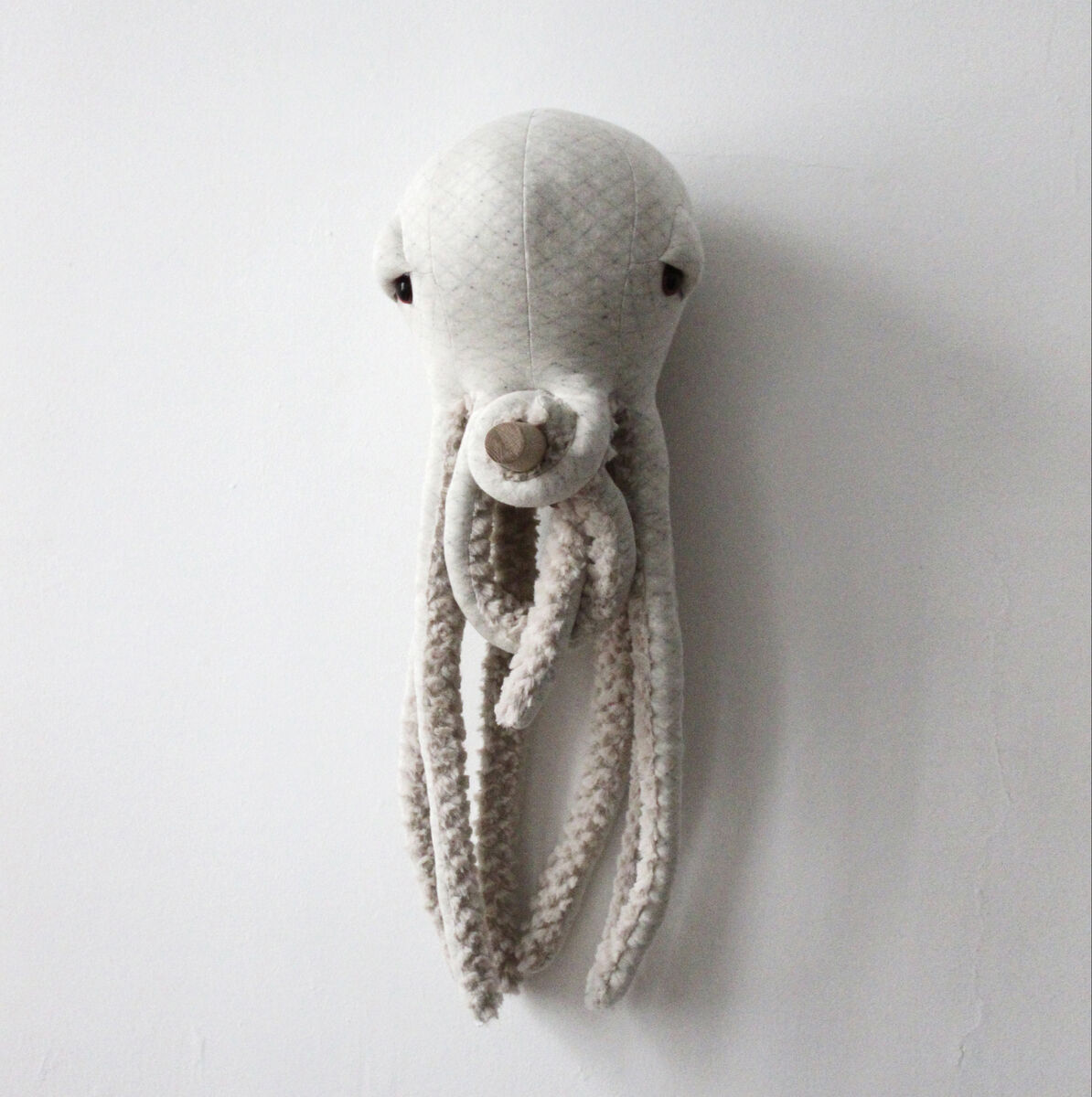 Ein Plüschtier in Form eines kleinen albino Oktopus, aufgehängt an eine weiße Wand. Das Kuscheltier ist handgefertigt und hat weiche, gestrickte Tentakeln.