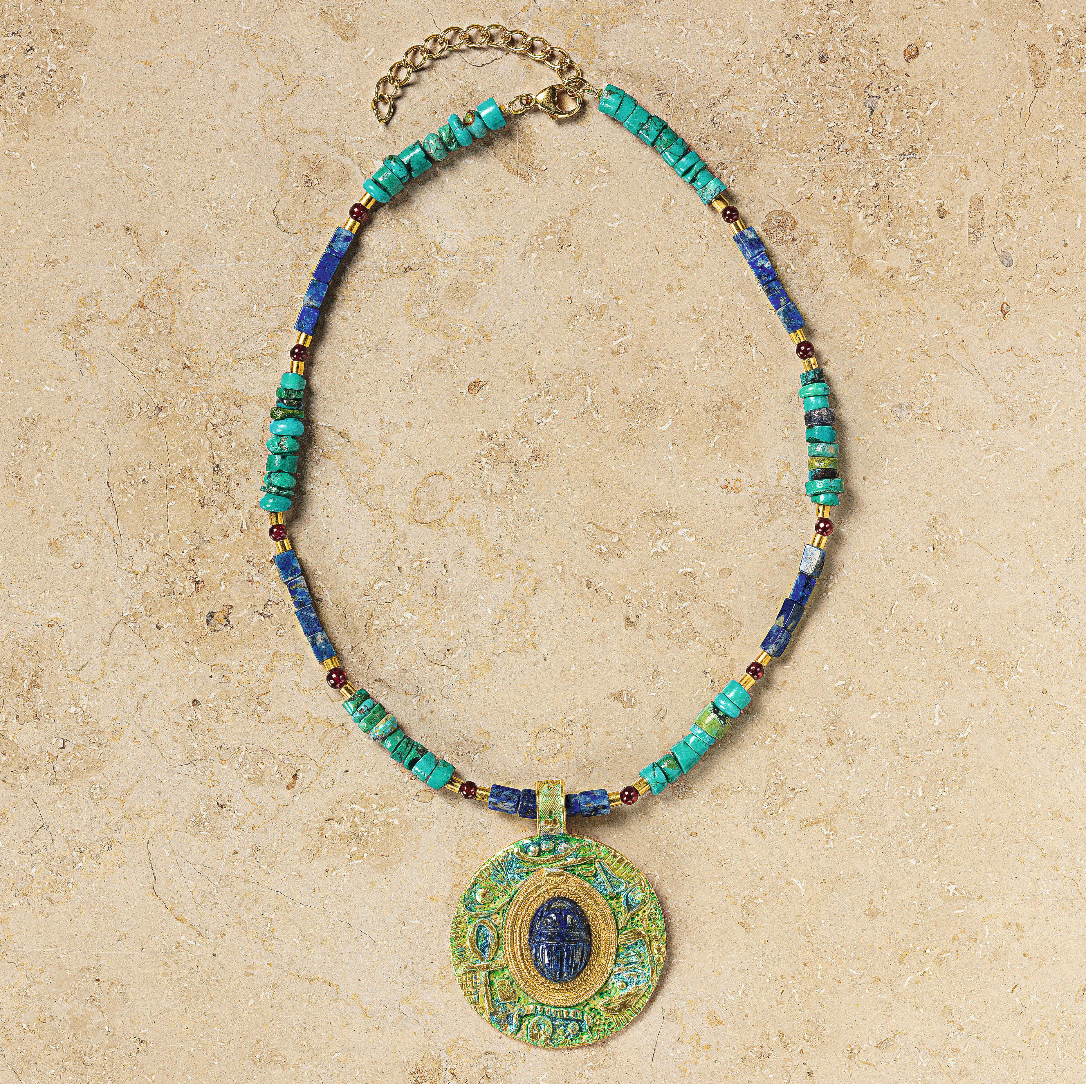 Das Collier »Scarabeo« von Petra Waszak zeigt einen zentralen Skarabäus-Anhänger aus tiefblauem Lapis in runder Form, eingefasst in vergoldetes 24-Karat-Messing. Die Kette setzt sich aus polierten Türkisen, Lapiskugeln und roten Granatperlen zusammen. Alle Elemente sind harmonisch aufeinander abgestimmt, die Oberfläche glänzt edel.