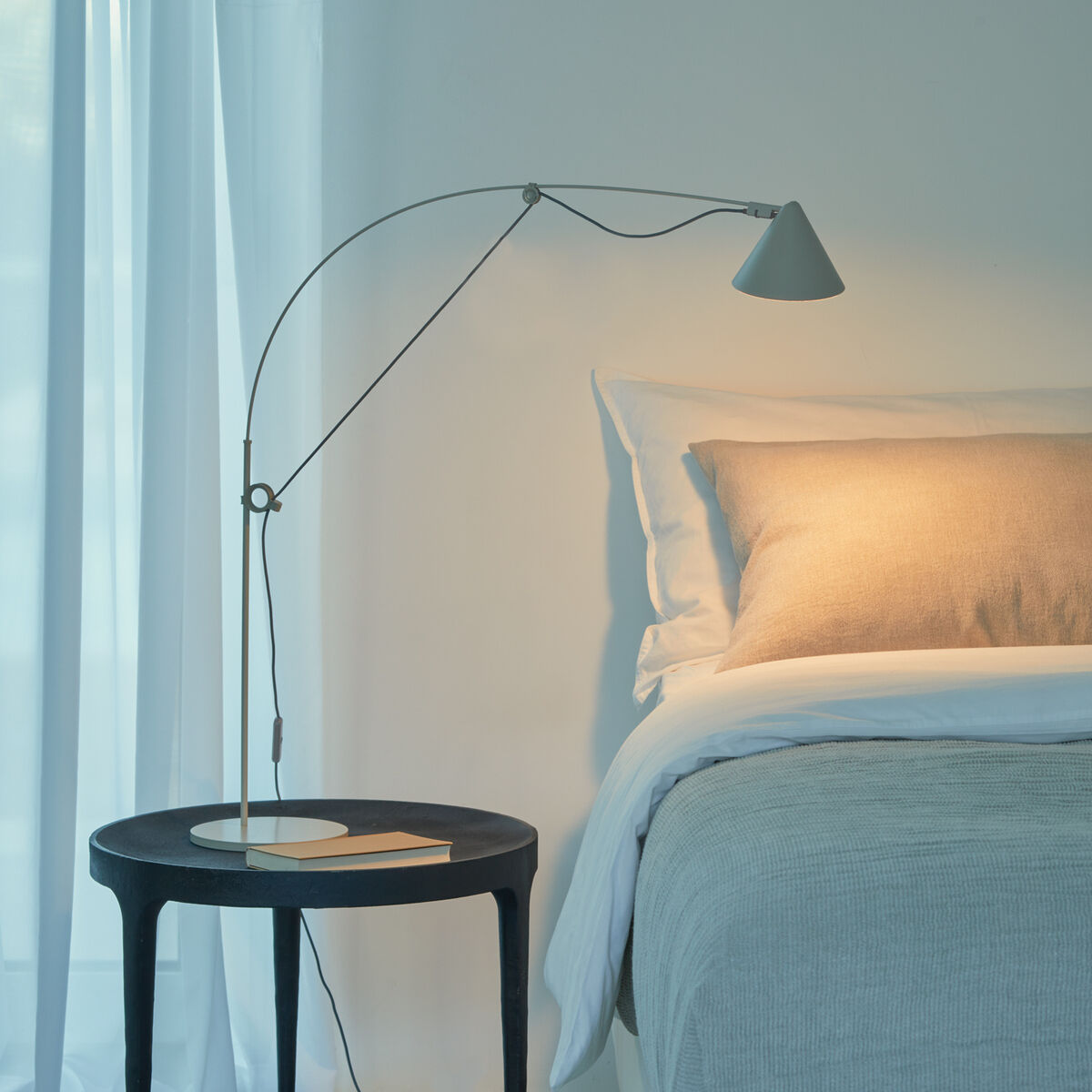 Moderne Tischlampe »AYNO« Silk auf einem schwarzen Beistelltisch neben einem Bett mit weißen Kissen und grauer Bettwäsche, umgeben von weißen Vorhängen in einem minimalistisch eingerichteten Schlafzimmer.