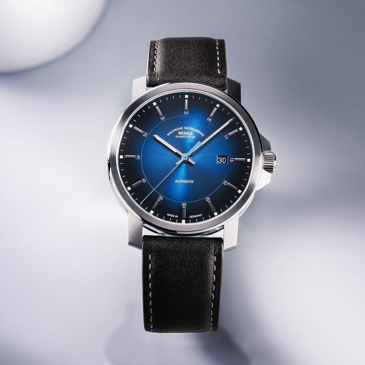 Die Automatikuhr »29er« Casual ZEIT-Sonderedition von Mühle-Glashütte ist auf einem hellen Untergrund edel arrangiert. Das Zifferblatt leuchtet intensiv blau mit silbernen Indizes und Zeigern, das Gehäuse ist aus Edelstahl, das Armband aus schwarzem Leder mit feiner Naht.