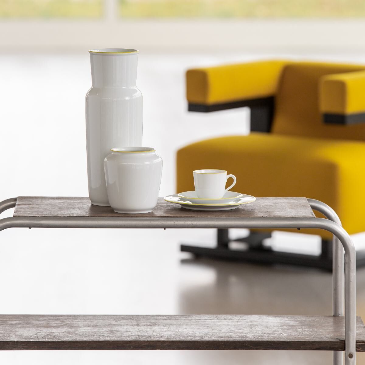 Die ZEIT-Sonderedition „Wagenfeld Vase 639/14“ von Fürstenberg zeigt eine schlanke, weiße Porzellanvase mit goldenen Akzenten. Daneben steht eine kleinere Tasse und eine leere Untertasse auf einem Tisch aus hellem Holz. Im Hintergrund ist ein gelber Stuhl sichtbar.