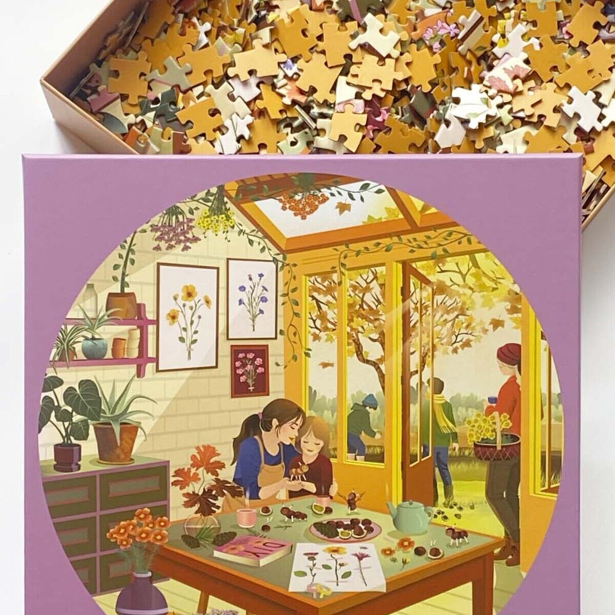 Die Puzzleverpackung zeigt eine herbstliche Szene in einem Wintergarten: Zwei Kinder und eine erwachsene Person basteln an einem Tisch mit Kastanien und Blättern. Draußen sammeln weitere Personen Blätter. Im Hintergrund sind herbstliche Bäume und gepresste Blätter als Dekoration zu sehen.