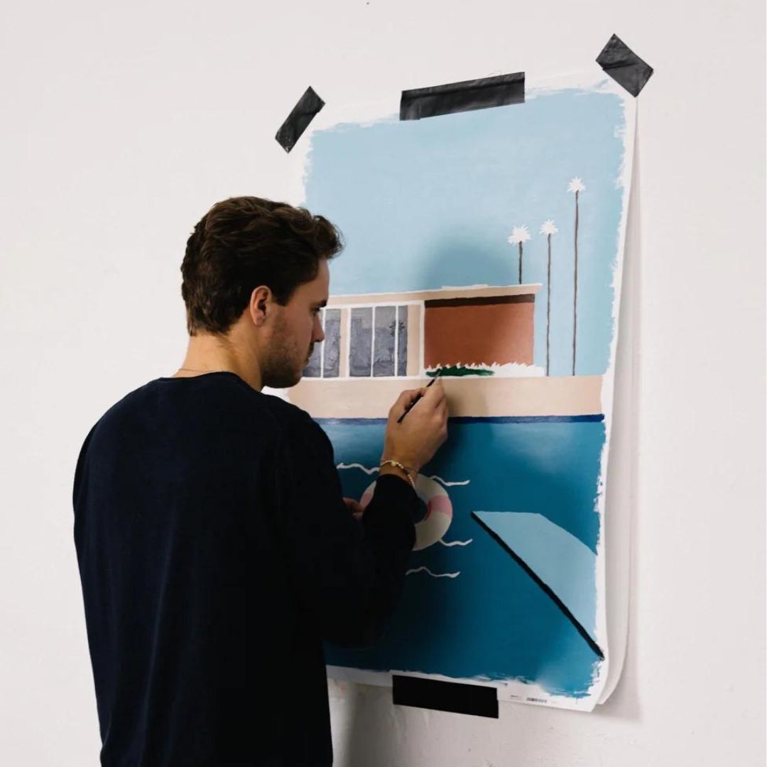 Ein Mann malt an einem 'Malen nach Zahlen'-Bild mit einem modernen Design auf weißem Hintergrund. Das Bild zeigt eine moderne Architektur mit einem Pool im Vordergrund. Schwarzweißes Klebeband hält das Papier an der Wand fest.