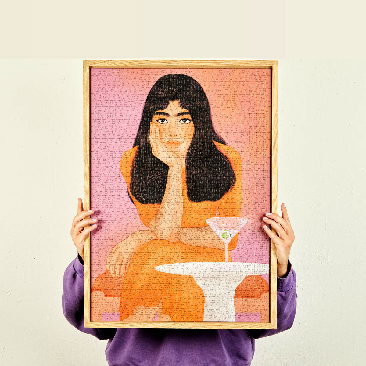 Puzzle »Staring into the Void« zeigt das Bild einer nachdenklichen Frau in einem orangefarbenen Outfit vor einem pinkfarbenen Hintergrund. Der Rahmen ist aus hellem Holz. Die Person im Bild hält ein Stück des Puzzles, das zum fertigen Kunstwerk gehört.