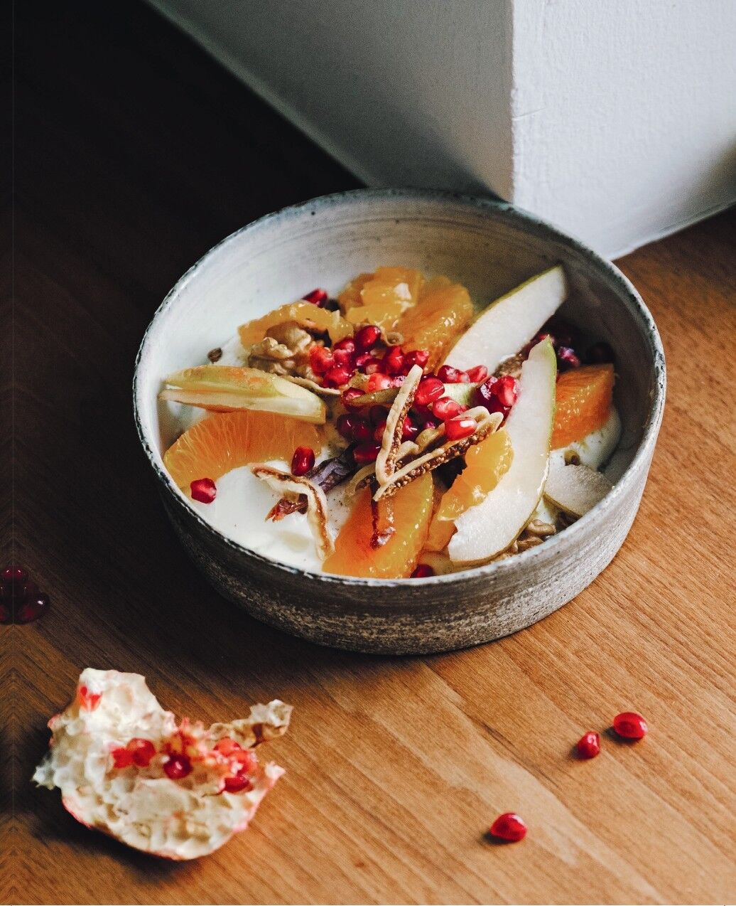 Eine Schüssel mit Joghurt, frischen Orangenscheiben, Granatapfelkernen, Nüssen und Müsli steht auf einem Holztisch. Daneben liegt eine angeknackte Granatapfelschale mit verstreuten Kernen. Das Bild vermittelt eine gemütliche Frühstücksatmosphäre in der Advents- und Weihnachtszeit.