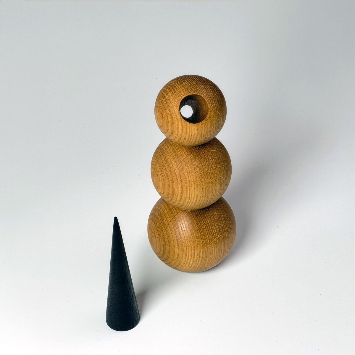 Minimalistische Holzfigur Schneemann »CLEAR« vor hellgrauem Hintergrund: Drei übereinandergestapelte, gedrechselte Kugeln aus warm honigbrauner Esche bilden den Körper. Der kleine Kopf ist ringförmig mit rundem Durchblick. Links steht separat ein schlanker, matt schwarzer Kegel als mögliche Nase.